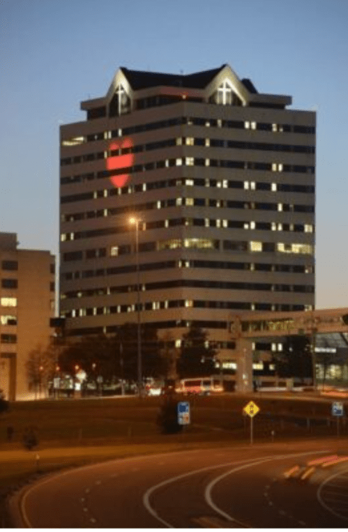 A tall building at dusk with a heart shape illuminated in its windows, surrounded by city lights and traffic.