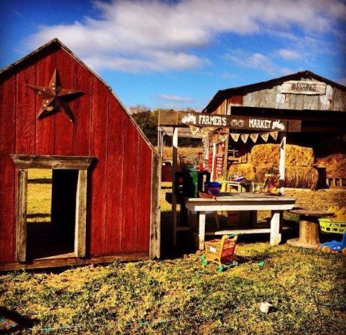 A rustic farmer's market with a red barn, wooden table, and play area surrounded by hay bales under a blue sky.
