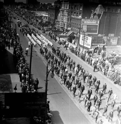 A historic black-and-white photo of a large parade with crowds lining the street, featuring marchers in uniform.