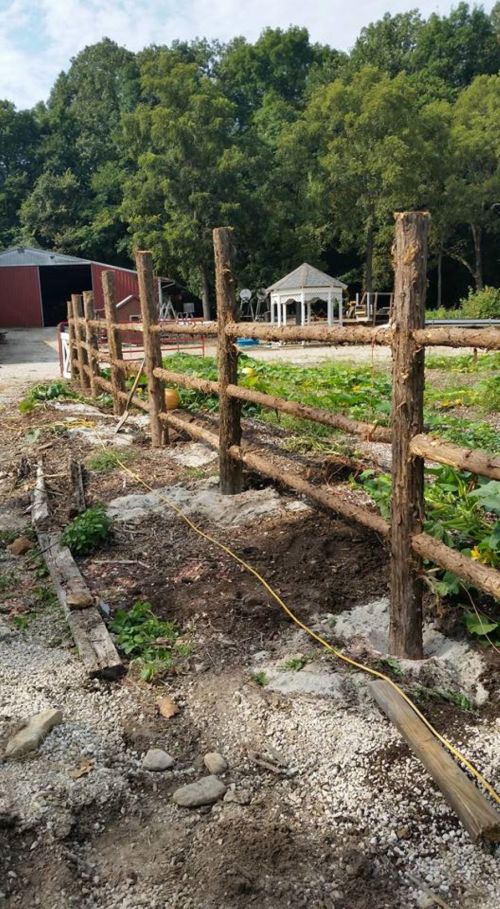 A rustic wooden fence lines a gravel path, with greenery and a gazebo visible in the background on a sunny day.