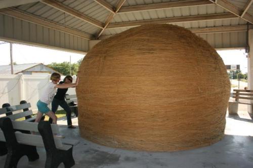 Two people push against a large, round structure made of straw in a covered area with benches.