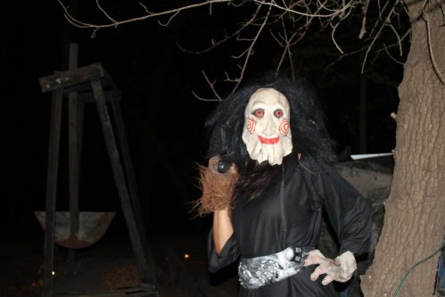 A figure in a creepy mask and black outfit poses in a dark setting, surrounded by eerie decorations.