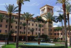 A hospital building with palm trees and a fountain in the foreground under a blue sky.