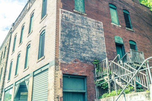 Historic brick building with faded signage and green shutters, featuring a staircase on the side.