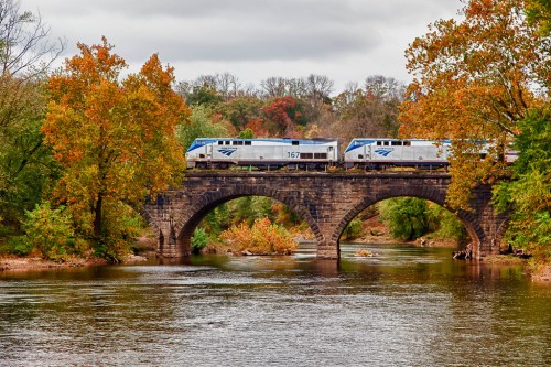 A train crosses a stone bridge over a river, surrounded by autumn-colored trees.