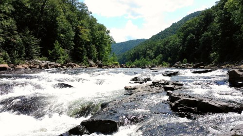 A scenic river flows over rocks, surrounded by lush green trees and mountains under a bright blue sky.