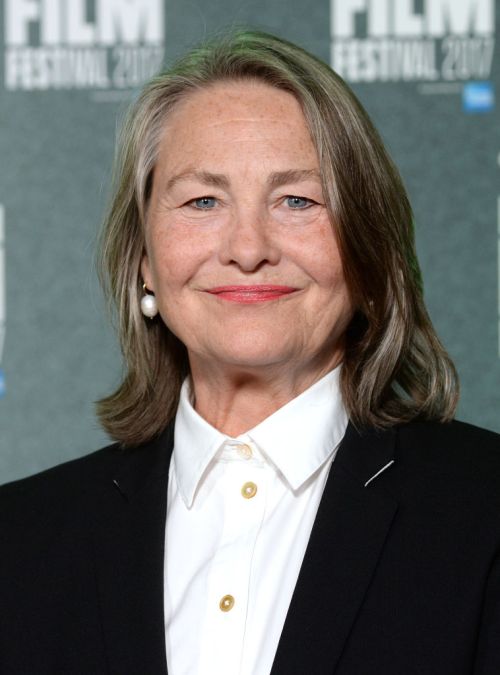A woman with long gray hair smiles, wearing a black suit and white shirt, at a film festival event backdrop.