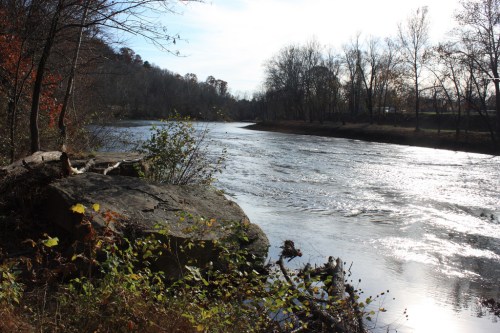 A serene river flows past rocky banks and trees, reflecting sunlight under a clear sky.