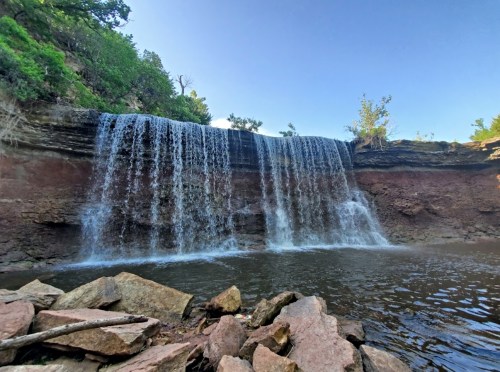 A serene waterfall cascading over rocky cliffs into a calm pool, surrounded by lush greenery and clear blue skies.