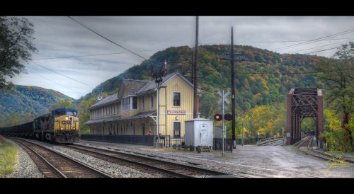 A train passes by a historic yellow station near a bridge, surrounded by colorful autumn hills under a cloudy sky.