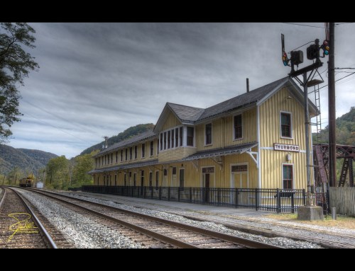 Historic yellow train station beside railway tracks, with a signal light and mountains in the background under a cloudy sky.