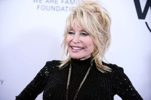 Dolly Parton smiles at an event, wearing a black sequined outfit and statement earrings against a white backdrop.