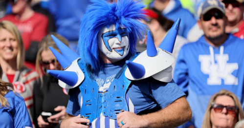 A fan with blue hair and face paint, wearing a blue shirt and spiked shoulder pads, stands out in a crowd.
