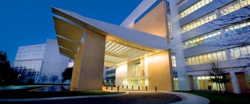 Modern building entrance with a large overhang, illuminated at dusk, surrounded by trees and a reflective pond.