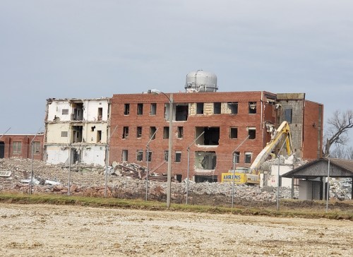 A partially demolished brick building with debris scattered around, surrounded by a fence and construction equipment nearby.