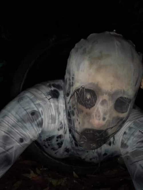 A creepy figure with a skull-like face emerges from a dark, shadowy background, partially obscured by a plastic covering.