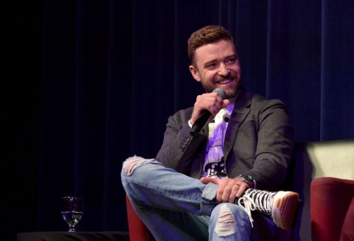 A smiling man in a blazer and jeans sits on stage, holding a microphone and engaging with the audience.