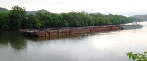 A barge navigates a calm river surrounded by lush green trees and rolling hills in the background.