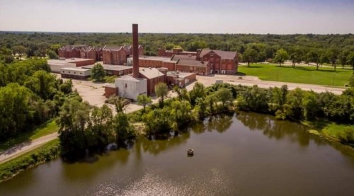 Aerial view of an old industrial complex beside a calm pond, surrounded by greenery and open fields.