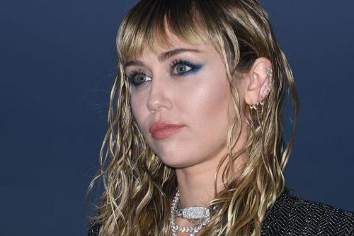 A close-up of a person with wet, wavy hair and bold blue eye makeup, wearing multiple earrings and a stylish outfit.