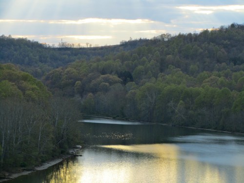 A serene river flows through lush green hills under a cloudy sky with rays of sunlight breaking through.