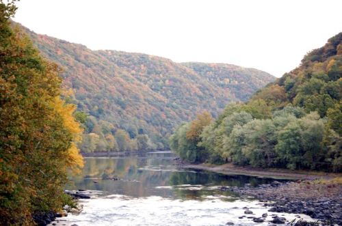 A serene river flows between colorful autumn hills, reflecting the vibrant foliage on the water's surface.