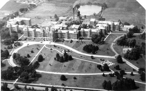 Aerial view of a large historic building surrounded by landscaped grounds and a pond in the background.