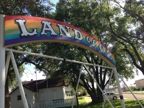A colorful archway reading "LAND of OZ" above a swing set, surrounded by trees and a small building in the background.