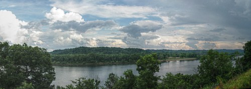 A panoramic view of a river surrounded by lush greenery and dramatic clouds in the sky.