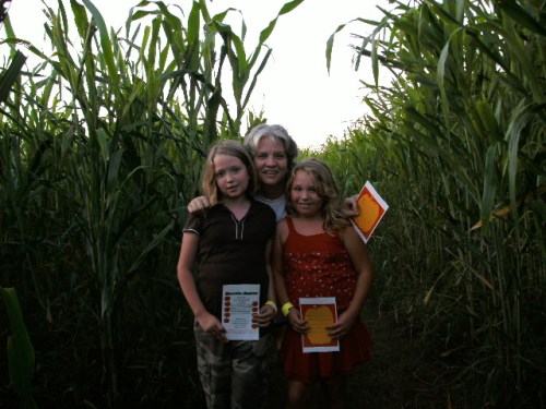 A woman and two girls stand in a cornfield, holding papers, surrounded by tall corn plants.