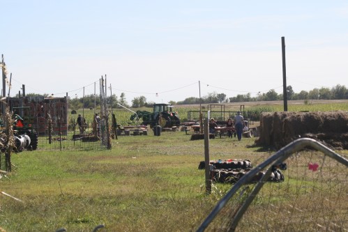 A rural scene with a tractor, people working, and farm equipment in a grassy field under a clear blue sky.