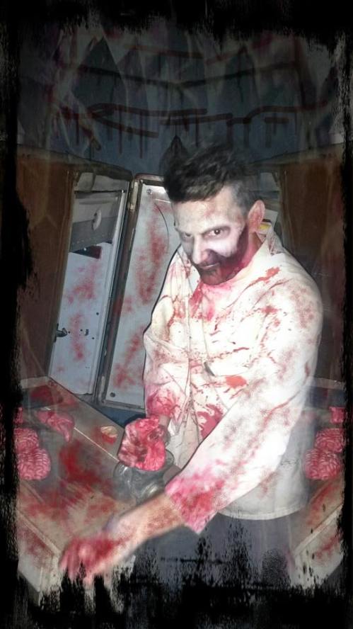 A bloodied figure in a tattered shirt stands over a table with brain-like objects, set in a dark, eerie environment.