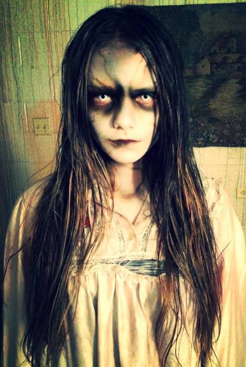 A ghostly figure with long, wet hair and haunting eyes, wearing a tattered white dress, stands in a dimly lit room.