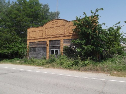 Abandoned brick building with boarded windows, surrounded by overgrown vegetation, along a rural road.