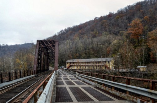 A rusted metal bridge spans a river, with a hillside of trees in autumn colors in the background.