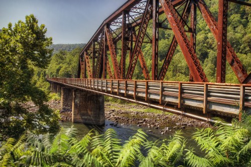 A rusted metal bridge spans a river, surrounded by lush greenery and rocky riverbanks under a clear blue sky.