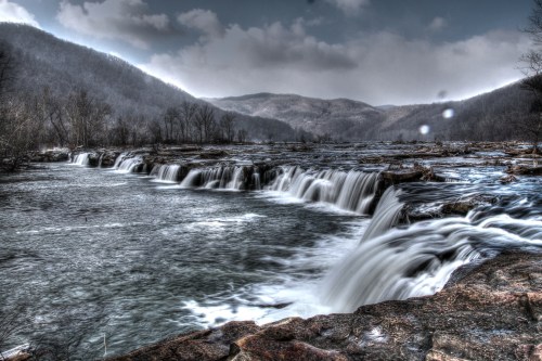 A serene landscape featuring cascading waterfalls and a river, surrounded by mountains under a cloudy sky.