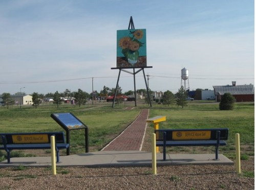 A park with two benches, a path, and a large painting of sunflowers on a stand in the background.