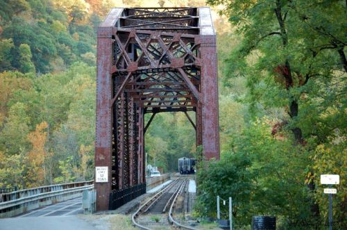 Rusty railroad bridge surrounded by colorful autumn foliage and a train track leading into the distance.