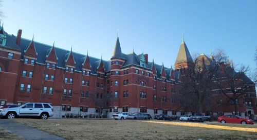Historic red brick building with pointed roofs and spires, surrounded by a grassy area and parked cars.