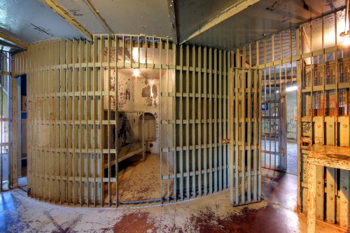 Interior of an old prison cell with barred doors and a dimly lit hallway, showcasing peeling paint and a stark atmosphere.