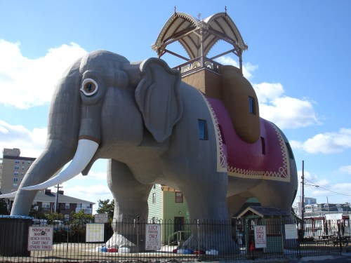 A large, whimsical elephant-shaped building with a saddle and a roof, surrounded by a fence and signs.