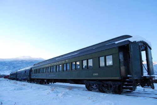 A vintage green train car on snowy tracks with mountains in the background under a clear blue sky.