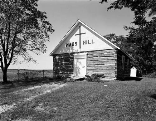 A small, rustic church building with a cross and "Mars Hill" sign, surrounded by trees and open fields.