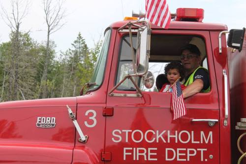 A firefighter and a child sit in the cab of a red fire truck, decorated with small American flags.