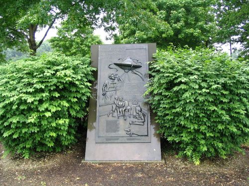 A bronze plaque depicting figures and text, surrounded by lush green bushes in a park setting.