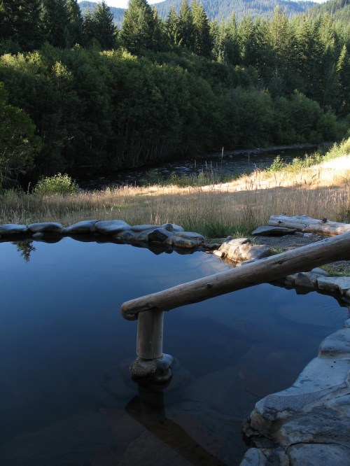 A serene hot spring surrounded by lush greenery and a river, with a wooden bridge leading over the water.