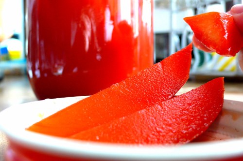 Bright red slices of fruit on a white plate, with a hand holding a piece, and a jar of red liquid in the background.