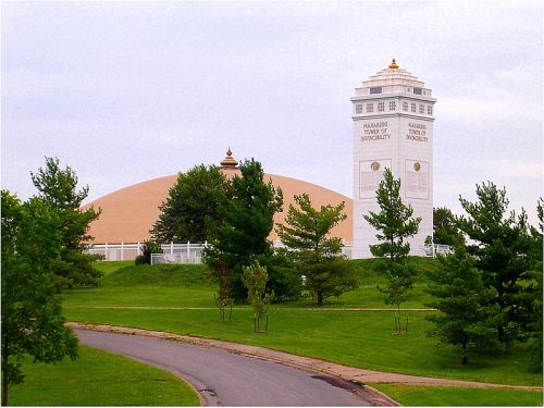 A large dome building and a white tower surrounded by green trees and a pathway under a cloudy sky.