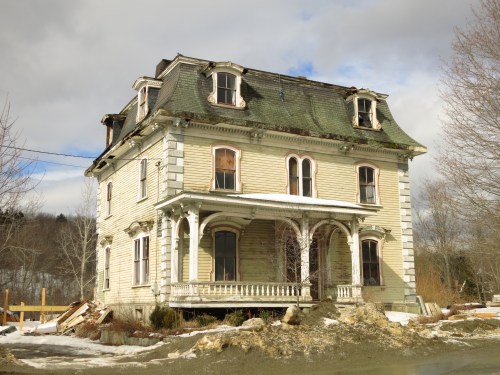 An old, dilapidated Victorian house with a green roof, broken windows, and overgrown landscaping.
